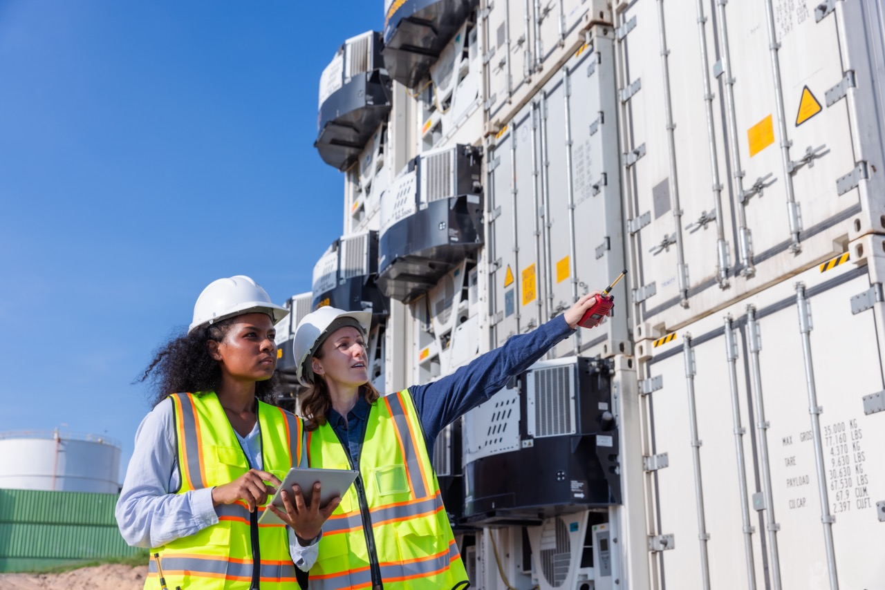 two workers in vests and hardhats looking at white refrigerated shipping containers in container yard